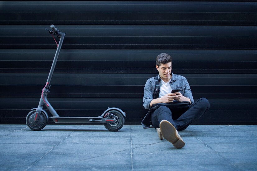 hipster-usando-telefono-inteligente-mientras-sentado-suelo-junto-su-scooter-electrico-contra-pared-n