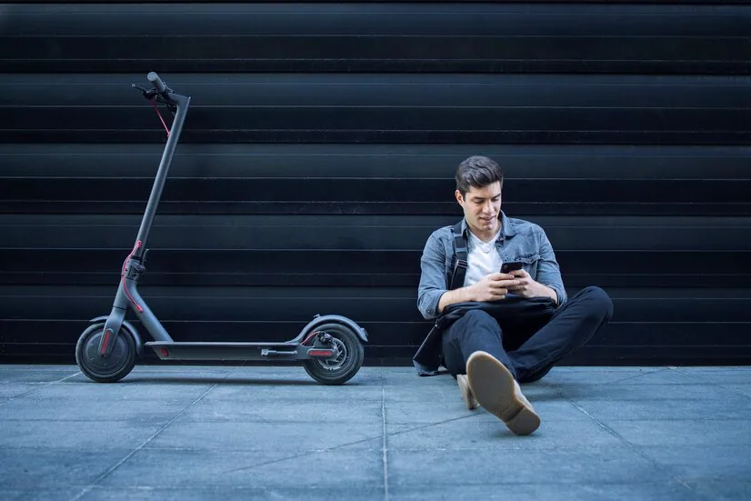 hipster-usando-telefono-inteligente-mientras-sentado-suelo-junto-su-scooter-electrico-contra-pared-n