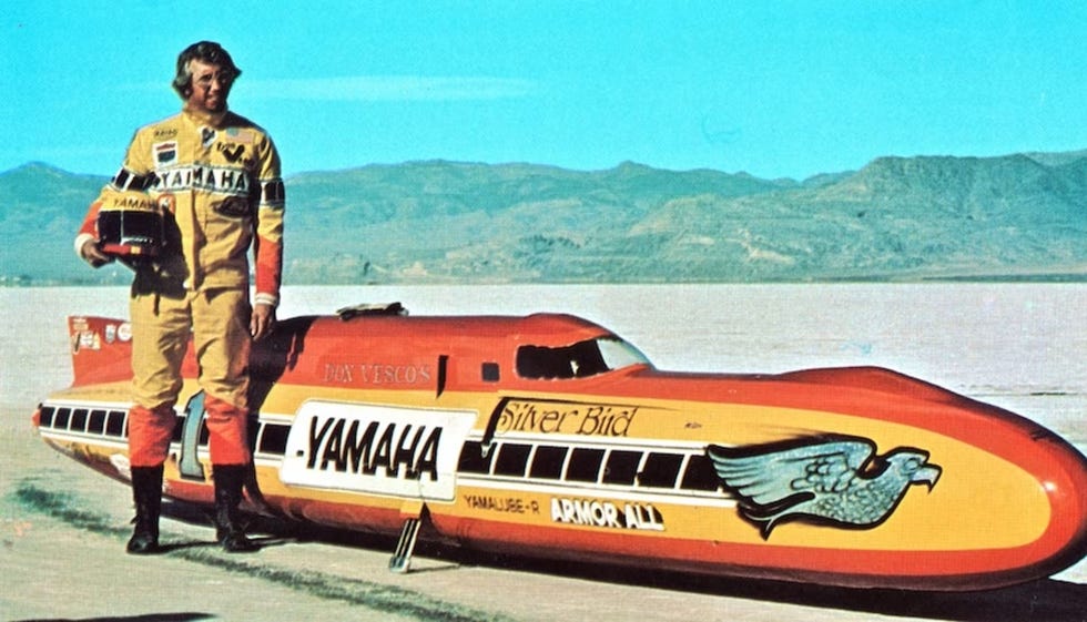 a man in a racing suit poses beside a streamlined vehicle on a flat desertlike surface under a clear blue sky